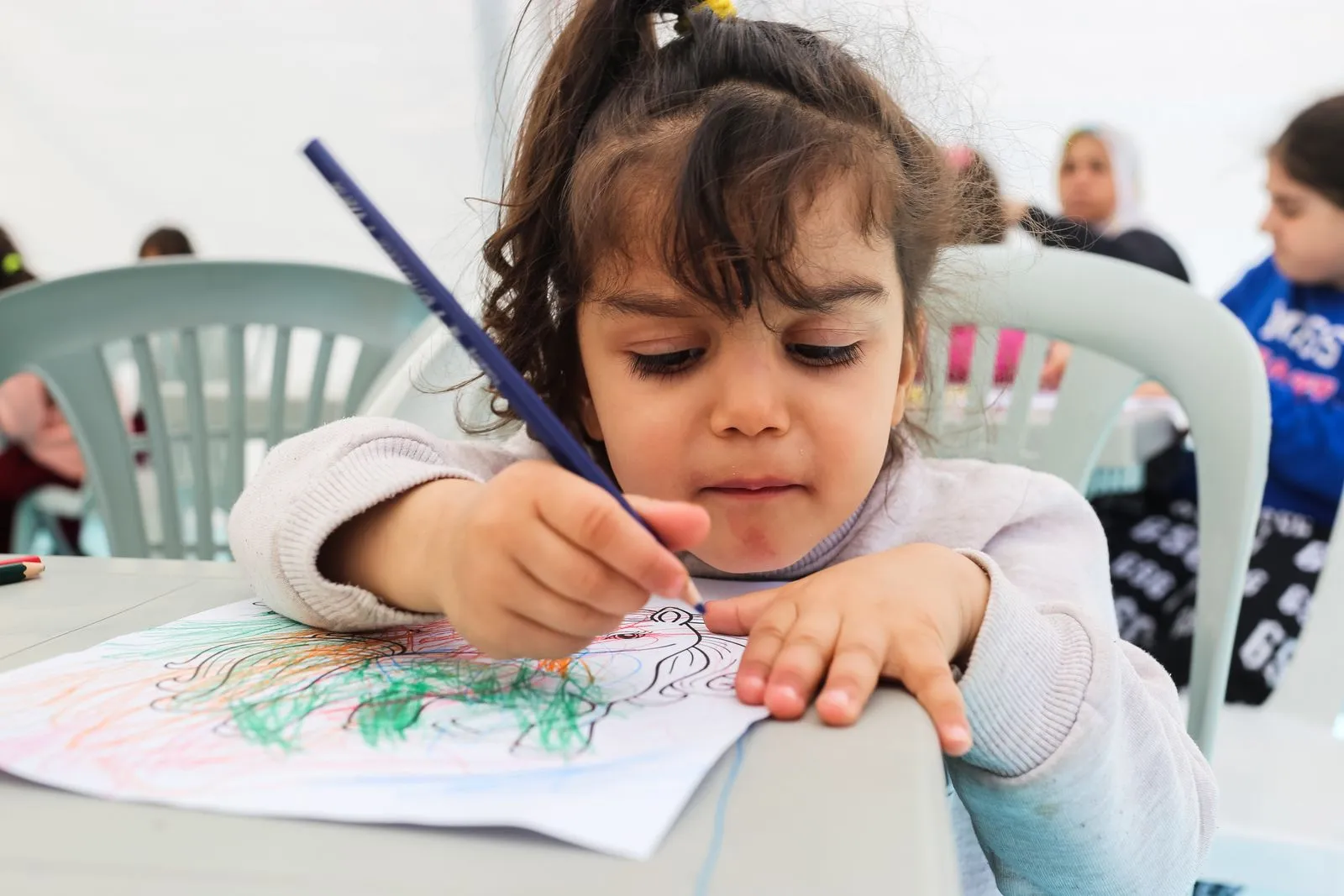 A young girl immersed in coloring a picture with crayons in a lively preschool setting.